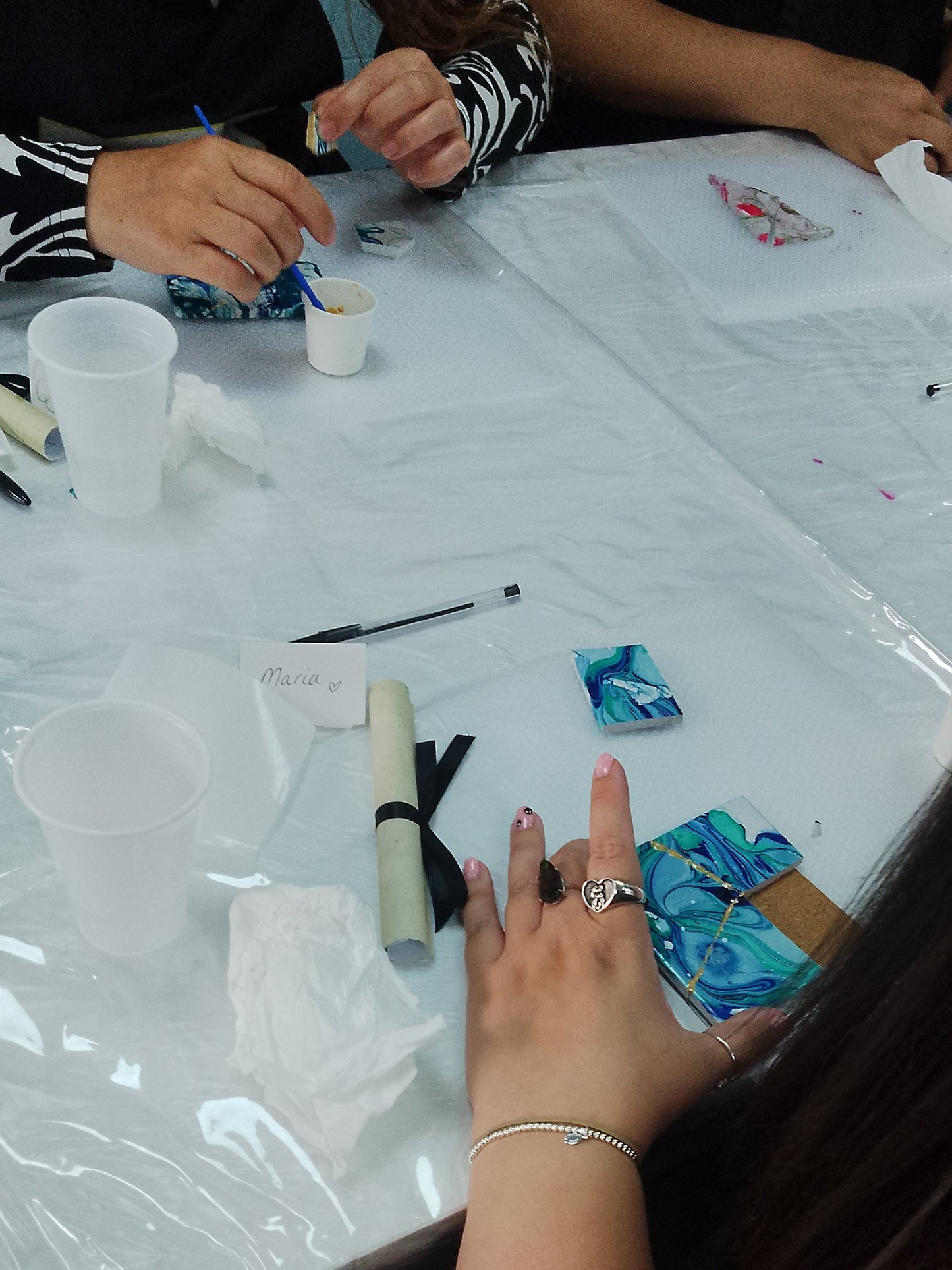 Person's hand with a ring on a table with art supplies and a small painting.