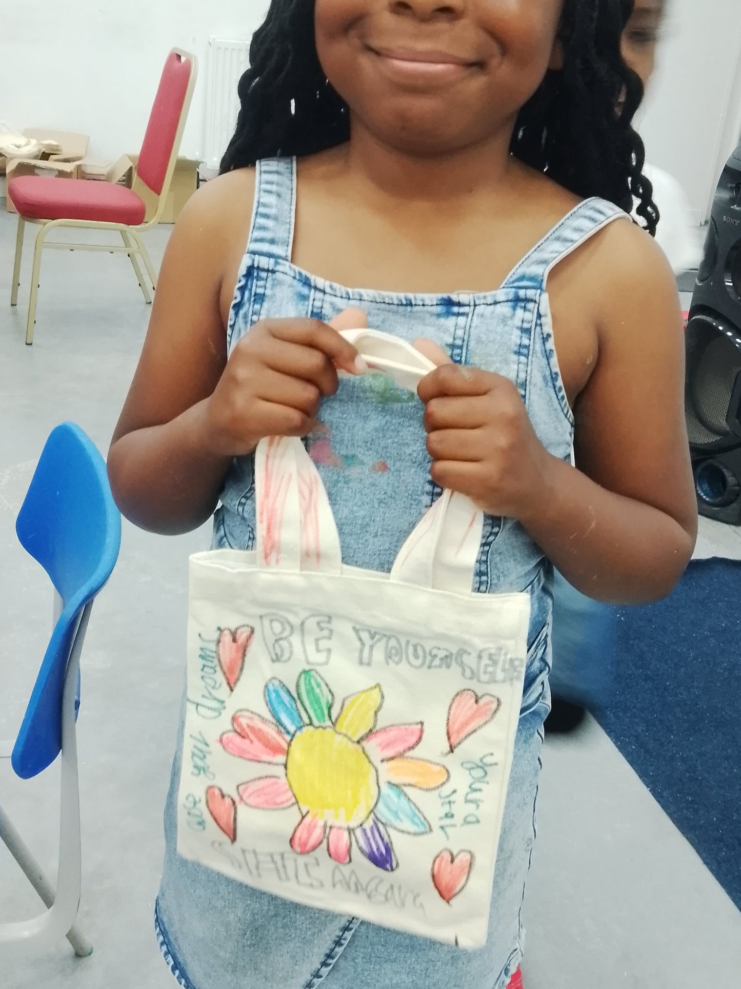 Young girl holding a decorated bag in an indoor setting with chairs and a speaker.