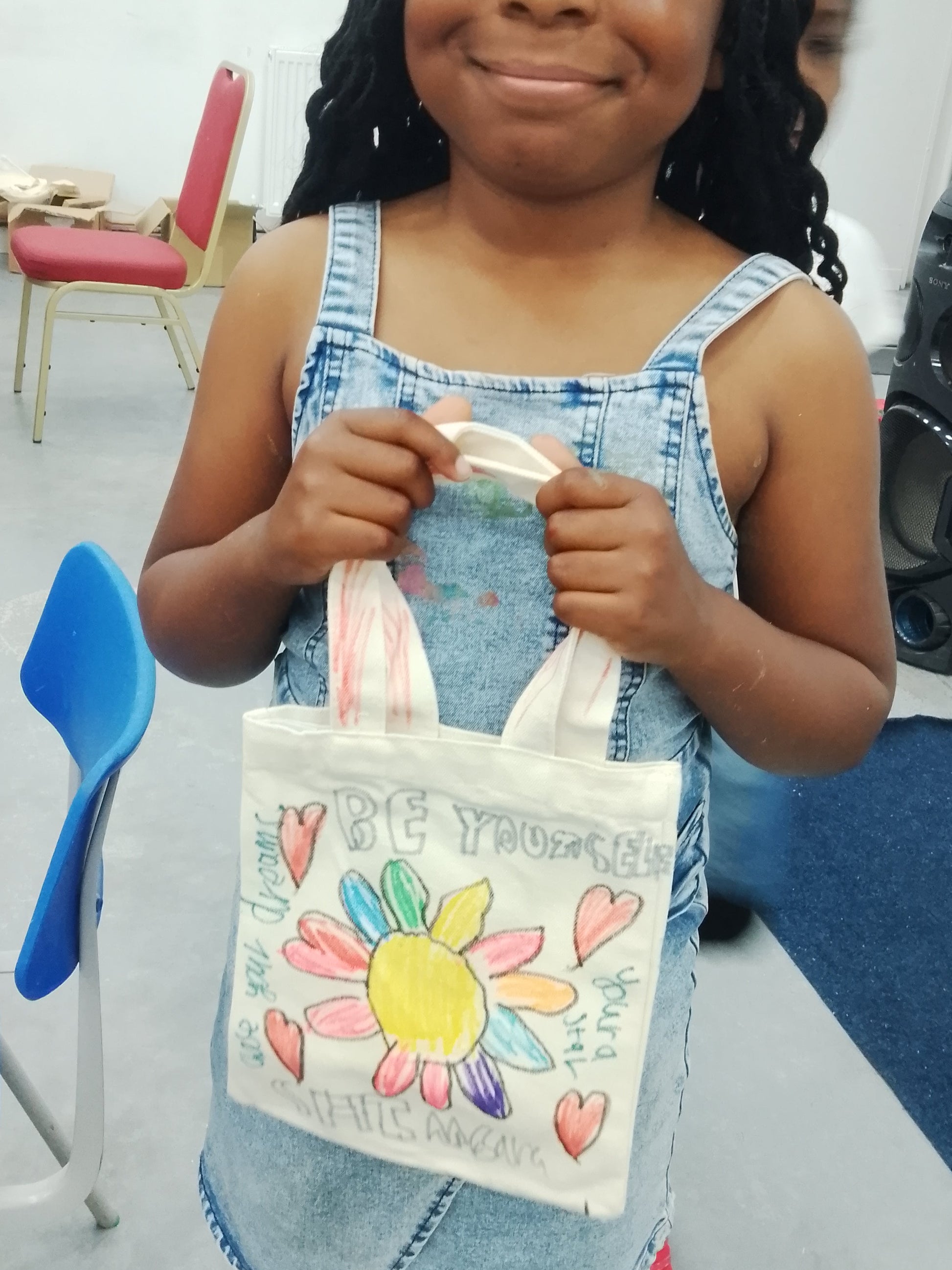 Young girl holding a decorated bag in an indoor setting with chairs and a speaker.